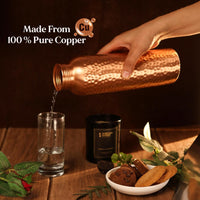 Copper bottle being poured into a glass with decorative elements on a wooden surface.