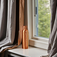 Copper water bottle on a windowsill with curtains and a view of greenery outside.