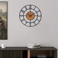 Decorative wall clock with Roman numeral design on a plain wall above a wooden shelf.